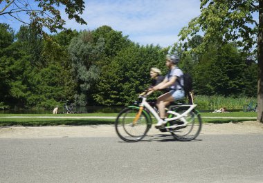 Vondelpark sıcak bir yaz gününde. İnsanlar güneşin altında ağaçların altında çimenlerin üzerinde dinleniyorlar. yolda iki Bisiklet sürme çift. Amsterdam, North-Holland, Hollanda 