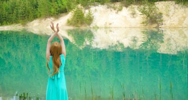 Sessiz sakin göl kenarında eli havada duran bir kadın. Güzellik, ayna efektli moda fotoğrafı. Genç kız gölün yanında tek başına duran uzun mavi bir elbise giyiyor. Hafta sonu açık hava. Arka plan