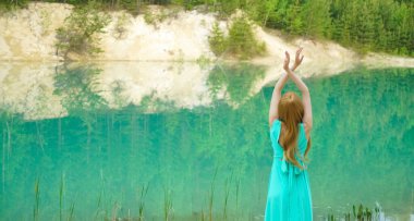 Sessiz sakin göl kenarında eli havada duran bir kadın. Güzellik, ayna efektli moda fotoğrafı. Genç kız gölün yanında tek başına duran uzun mavi bir elbise giyiyor. Hafta sonu açık hava. Arka plan