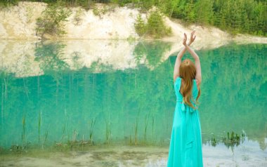 Sessiz sakin göl kenarında eli havada duran bir kadın. Güzellik, ayna efektli moda fotoğrafı. Genç kız gölün yanında tek başına duran uzun mavi bir elbise giyiyor. Hafta sonu açık hava. Arka plan