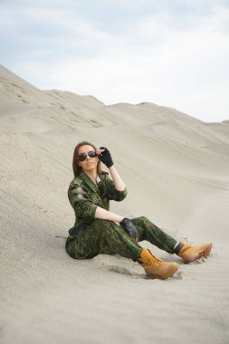 Üniformalı bir kadın kum tepesinde silahsız. Tatlının arka planında askeri kostümlü güzel bir kadın var. Rus haki üniforması.