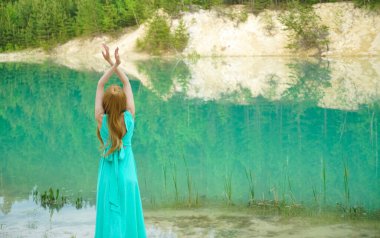 Sessiz sakin göl kenarında eli havada duran bir kadın. Güzellik, ayna efektli moda fotoğrafı. Genç kız gölün yanında tek başına duran uzun mavi bir elbise giyiyor. Hafta sonu açık hava. Arka plan