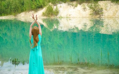 Sessiz sakin göl kenarında eli havada duran bir kadın. Güzellik, ayna efektli moda fotoğrafı. Genç kız gölün yanında tek başına duran uzun mavi bir elbise giyiyor. Hafta sonu açık hava. Arka plan