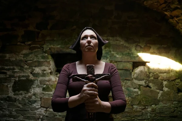 Religious life concept -  nun prays in the archival dark medieval monastery. middle age woman  holding a stone cross on stoned wall background
