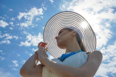 Parlak güneş ışığı, geniş kenarlı şapkalı, mavi bulutlu gökyüzü arka planlı güzel genç kızın yüzünde parlıyor. Düşük açı atışı