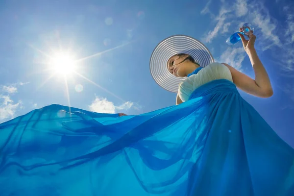 ローアングル撮影 青いドレスと帽子を身に着けている魅力的な若い女性 青い空の背景に青いシャンパンを飲みます — ストック写真