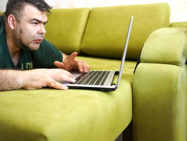 Yeşil kanepede uzanan ve dizüstü bilgisayarda çalışan meşgul bir adam..