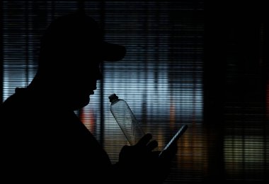 Şeffaf camlara karşı plastik şişeden içen bir adamın silüeti. Erkek, su şişesi içiyor. Erkek cep telefonu kullanıyor. gece sahnesi.