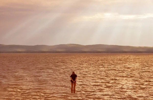 Woman seen from behind entering the sea clothed. Standing in clear beautiful sea in water, beach. Mountains and endless sea in the distance. rays of the sun through dramatic clouds