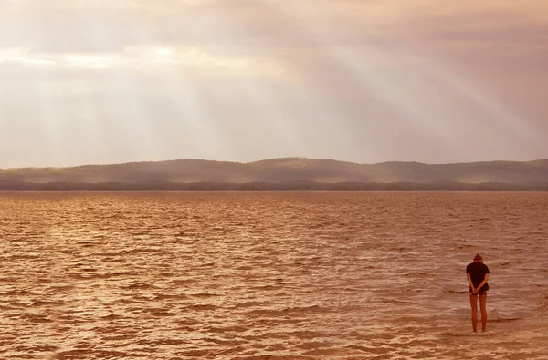 Woman seen from behind entering the sea clothed. Standing in clear beautiful sea in water, beach. Mountains and endless sea in the distance. rays of the sun through dramatic clouds