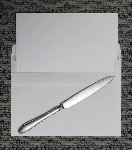 Blank paper with knife and letter lies on pattern. A top down view of a white envelope and a letter opener on a paper surface.