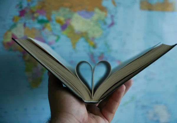 pages of a book curved into a heart shape. globe map on background. male hand holding book