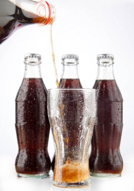 Pouring Cola of plastic bottle in transparent glass with foam on three full flask isolated on white background. clipart