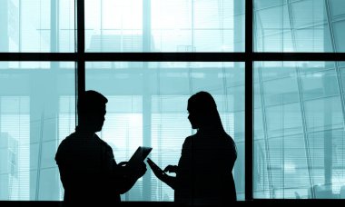 İki iş adamının siluetleri. Adam ve kadın ofis içi, adam tabletinin ekranında iş arkadaşına bilgi gösteriyor, kocaman pencereler. 