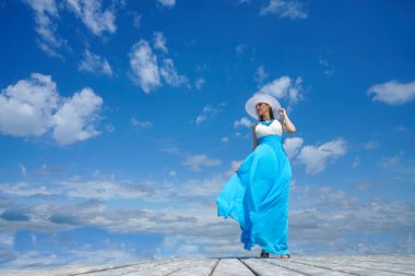 Beyaz ve mavi elbiseli kız ahşap bir iskelede duruyor. Mavi bulutlu bir gökyüzünün arka planına karşı. Rüzgarlı bir hava. Yüzünde ışık ve gölgeler.