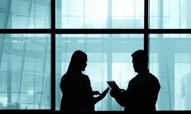 İki iş adamının siluetleri. Adam ve kadın ofis içi, adam tabletinin ekranında iş arkadaşına bilgi gösteriyor, kocaman pencereler. 