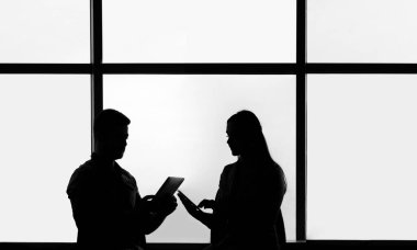 İki iş adamının siluetleri. Adam ve kadın ofis içi, adam tabletinin ekranında iş arkadaşına bilgi gösteriyor, kocaman pencereler. 