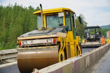Otoyol onarımı, asfalt sıkıştırıcı yaz gününde mavi gökyüzünde ve yeşil çam ormanlarında arka planda, yol inşa makinelerinde