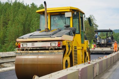 Otoyol onarımı, asfalt sıkıştırıcı yaz gününde mavi gökyüzünde ve yeşil çam ormanlarında arka planda, yol inşa makinelerinde