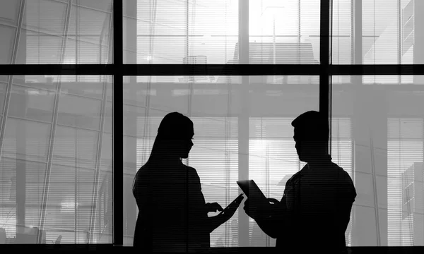 Silhouettes of 2 two business people man and woma in office interior, man showing information on screen of his tablet to his colleague, huge windows