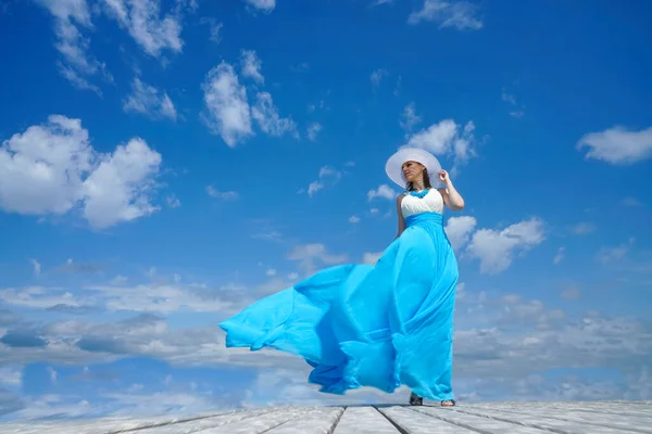 白と青のドレスの少女は木製の桟橋に立っている 青空を背景に 風の強い天気 顔の光と影 — ストック写真