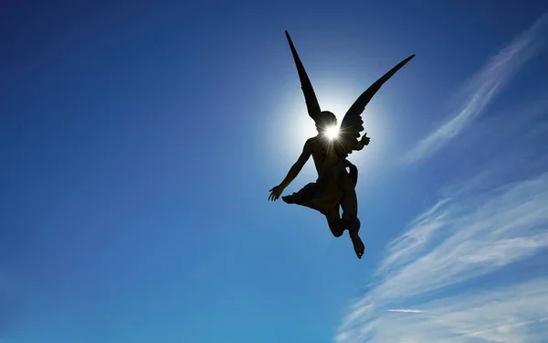 Low angle backlight portrait over a religious statue of an angel brandishing a christian catholic cross,  man with wings in his back and blue sunny sky.