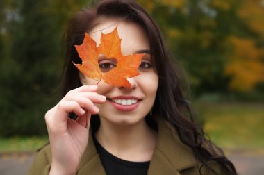 Autumn woman holding yellow maple leaves and hiding her eye. Cute female model looks out of yellow leaves. Happy  smiling blond teen girl with autumn leaf in the heart form.