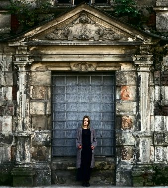 Beton duvarın önünde güzel bir kız. Moda. Tarz sahibi siyah elbiseli ve gri ceketli güzel bir kadın terk edilmiş tarihi bir evin girişinde duruyor.