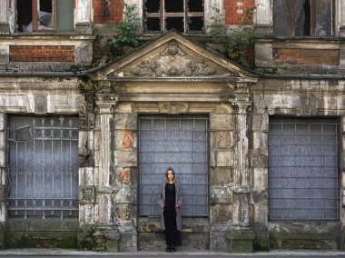 Beton duvarın önünde güzel bir kız. Moda. Tarz sahibi siyah elbiseli ve gri ceketli güzel bir kadın terk edilmiş tarihi bir evin girişinde duruyor.