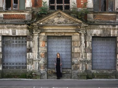 Beton duvarın önünde güzel bir kız. Moda. Tarz sahibi siyah elbiseli ve gri ceketli güzel bir kadın terk edilmiş tarihi bir evin girişinde duruyor.