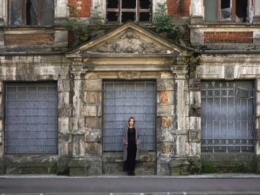 Beton duvarın önünde güzel bir kız. Moda. Tarz sahibi siyah elbiseli ve gri ceketli güzel bir kadın terk edilmiş tarihi bir evin girişinde duruyor.