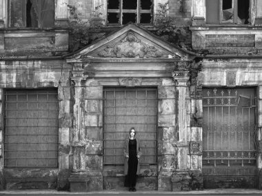 Beton duvarın önünde güzel bir kız. Moda. Tarz sahibi siyah elbiseli ve gri ceketli güzel bir kadın terk edilmiş tarihi bir evin girişinde duruyor.