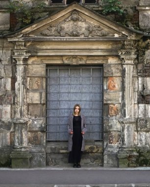 Beton duvarın önünde güzel bir kız. Moda. Tarz sahibi siyah elbiseli ve gri ceketli güzel bir kadın terk edilmiş tarihi bir evin girişinde duruyor.