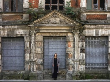 Beton duvarın önünde güzel bir kız. Moda. Tarz sahibi siyah elbiseli ve gri ceketli güzel bir kadın terk edilmiş tarihi bir evin girişinde duruyor.