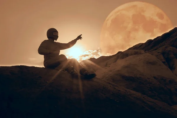 spaceman pointing at beautiful sky with stars while sitting on top of mountain. Mission specialist astronaut wearing white space suit with helmet. Concept of space travel and night sky. Side view