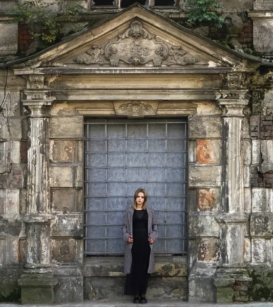 Beton duvarın önünde güzel bir kız. Moda. Tarz sahibi siyah elbiseli ve gri ceketli güzel bir kadın terk edilmiş tarihi bir evin girişinde duruyor.
