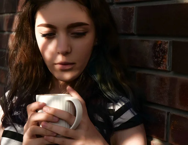 Sabah kahvesiyle tuğla duvarın yanında duran çok güzel bir kız. Güneş ışığı ve evin gölgesi. Kadın aşağı bakıyor..