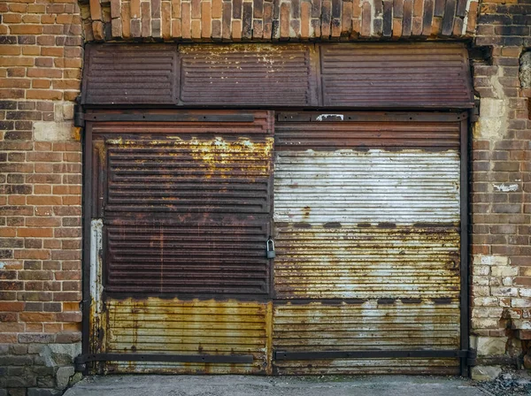 Eski bir garajın ön cephesi kırmızı tuğladan yapılmış büyük paslı kapılı yeşil çimlerde. Ön manzara. asma kilit