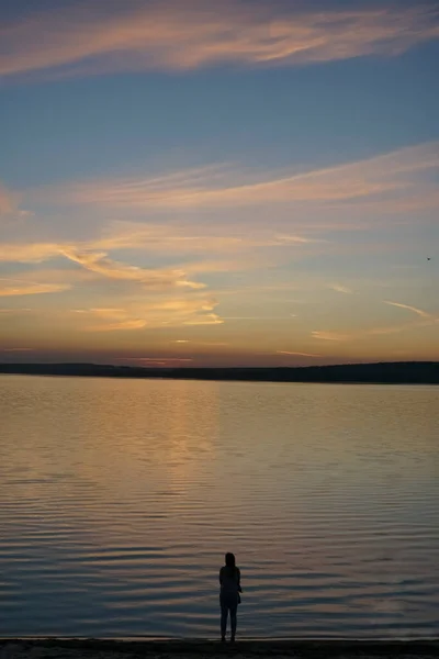 silhouette of woman standing near the water looking at the sunset across the lake. woman wearing  jeans and jacket  standing in sea water at the beach enjoying view of rising sun in early  morning