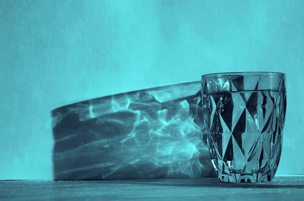 Water glass with strong shadow on  sunny wall  background. blue faceted glass. pure drinking water. sunlight.