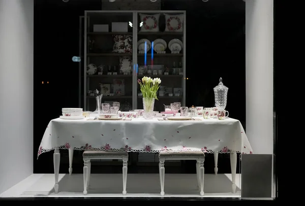 table with white plates, crystal glasses, linen napkins, cutlery and tulip flowers on luxurious tablecloth. shop window. night scene