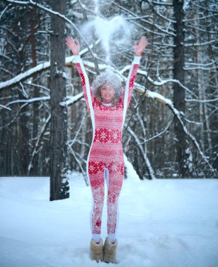 Sarışın kadın ormanda havaya kar fırlatıyor. Kız kırmızı atkı takıyor, gri kürk şapka takıyor. Hava güzel ve kar yağıyor. Tam boy kadın.