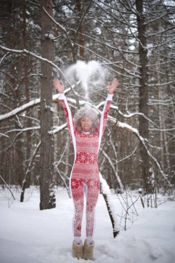 Sarışın kadın ormanda havaya kar fırlatıyor. Kız kırmızı atkı takıyor, gri kürk şapka takıyor. Hava güzel ve kar yağıyor. Tam boy kadın.