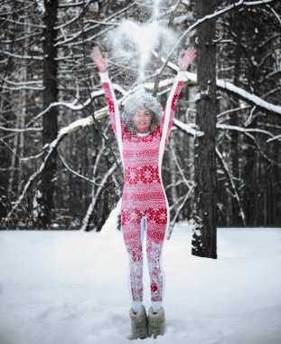 Sarışın kadın ormanda havaya kar fırlatıyor. Kız kırmızı atkı takıyor, gri kürk şapka takıyor. Hava güzel ve kar yağıyor. Tam boy kadın.