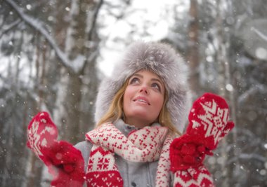 Forest Park 'taki sarışın kadın portresi. Kürk kürk şapkalı, kırmızı eldivenli, beyaz eşarplı güzel bir kız. Kış modası. Mavi gözler. Arka planda karlı ağaçlar var. Yukarı bakıyorum.