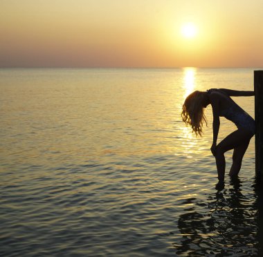 Deniz kıyısında duran bir kadın silueti. Güneş ışığı. Kız güneşli günbatımının tadını çıkarıyor. Sarı uzun saçlı..