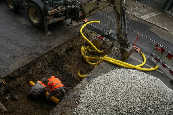 掘削機と2人の掘削機が公共道路を横断する排水管を敷設するために溝を掘る 地下の段ボールパイプ敷設 — ストック写真
