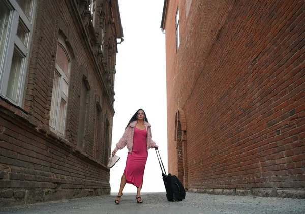 女人穿粉红色长裙 手握粉色运动鞋 提着行李走路的女人 带轮子的手提箱轻装旅行 一个女孩在古老的砖房之间沿着一条狭窄的街道走着 — 图库照片
