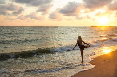 Gün batımı kumsalında antrenman yapmadan önce bacağını ısıtan genç bir kadının silueti. Kum ve deniz suyuna yansıyan güneş ışığı. sağlıklı yaşam tarzı