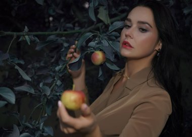 Sonbahar elbiseli kadın elma ağacının altında duruyor ve kırmızı elmaya bakıyor. Kadın elinde kırmızı ve yeşil elma. 